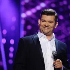 Zenek Martyniuk w czarnym garniturze i białej koszuli trzyma mikrofon na tle fioletowych świateł.
