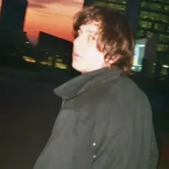 Francesco De Leo, con i capelli castani mossi, indossa un cappotto scuro. Sullo sfondo, un tramonto arancione illumina i profili degli edifici urbani.