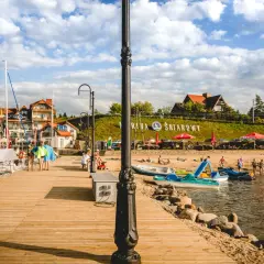 Letnia przygoda w krainie tysiąca jezior. Drewniane molo, żaglówki, plaża z ludźmi i budynki nad jeziorem.
