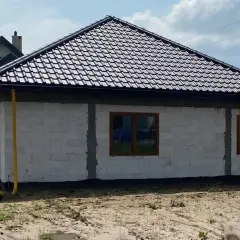 Dom w budowie z żółtymi rurami odprowadzającymi wodę z rynien. Widać białe bloczki, brązowe okna i dach pokryty ciemną dachówką.