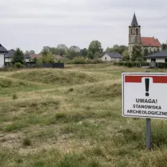 Znak "Uwaga! Stanowiska archeologiczne" na tle zabudowań i kościoła.