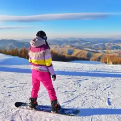 Snowboardzistka w różowych spodniach i kolorowej kurtce na tle górskiego krajobrazu.