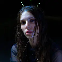 Ginevra, la giovane cantante, con capelli bagnati e sguardo intenso, si prepara per un'esibizione notturna sotto luci soffuse.