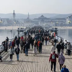 Tłum ludzi spaceruje po najdłuższym molo w Polsce, podziwiając widok na morze i nadmorskie budynki.