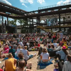 Tłum ludzi na festiwalu muzycznym, siedzących na ziemi i słuchających koncertu. W tle widać scenę i wielki ekran z występującymi artystami. To część owf lineup.