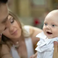 Un bébé souriant en tenue blanche, entouré de ses parents. Un moment précieux pour un baptême civil, depuis quand cette tradition existe-t-elle ?