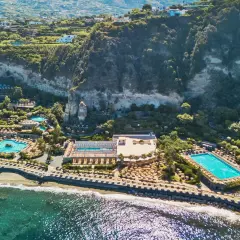 Vista aerea di lussuosi complessi termali a Ischia, con piscine scintillanti, lettini e vegetazione rigogliosa che si affacciano sul mare.