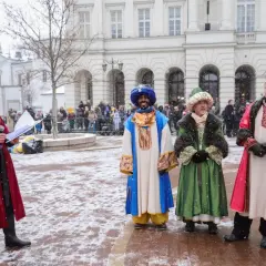 Król Melchior w niebieskim stroju z turbanem, obok innych postaci w barwnych szatach, podczas zimowego wydarzenia.