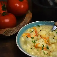 Pyszna zupa kalafiorowa z makaronem, ozdobiona natką pietruszki. Obok miski leżą świeże pomidory.