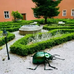 Ogród klasztorny z rzeźbą zielonego chrząszcza, studnią i żywopłotami.