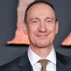 Uśmiechnięty David Thewlis w garniturze i krawacie, z niebieskimi oczami, na tle rozmytych, pomarańczowych kształtów.