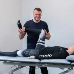 Fizjoterapeuta pomaga pacjentce w ćwiczeniach rehabilitacja kolana po urazie. Kobieta leży na kozetce, mężczyzna delikatnie zgina jej nogę.
