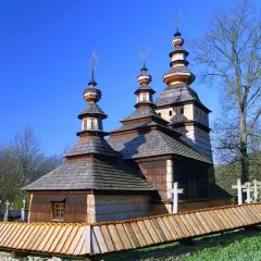 Drewniana cerkiew z trzema kopułami, otoczona płotem, w jednej z malowniczych miejscowości Beskidu Niskiego.