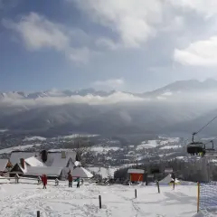 Zimowy krajobraz z wyciągiem krzesełkowym, ośnieżonymi domkami i Tatrami w tle.