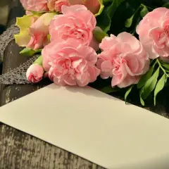Bukiet różowych kwiatów, koronka i pusta kartka na drewnianym tle.