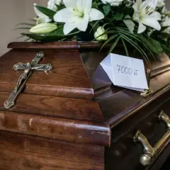 Drewniana trumna z krzyżem i kwiatami. Na wieczku karteczka z napisem "7000 zł", sugerująca, ile kosztuje pogrzeb katolicki.