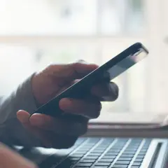 Dłoń trzyma telefon, który nie widzi laptop. Klawiatura komputera widoczna w zbliżeniu.