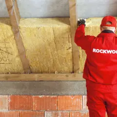 Pracownik w czerwonym stroju Rockwool montuje izolację z wełny mineralnej, tworząc zabudowę murłaty od zewnątrz.