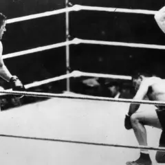 Jack Dempsey vs Gene Tunney in the ring. Dempsey stands, while Tunney is on one knee, with the referee looking on.