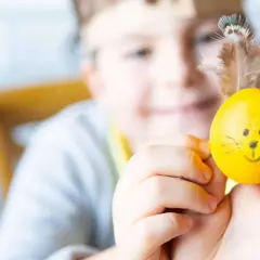 Ein Kind hält ein gelbes Ei mit lustigen Ostermotiv-Gesicht und Federn als Ohren.