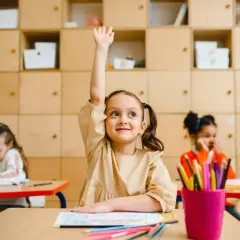 Dziewczynka z podniesioną ręką siedzi przy biurku w klasie, obok inne dzieci.
