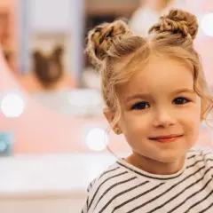 Urocza dziewczynka z blond włosami upiętymi w dwa kucyki. Jej fryzura to idealne włosy dla dziewczyn, które chcą wyglądać modnie i dziewczęco.