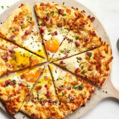 Kawałki pizzy carbonara z jajkiem sadzonym, boczkiem i szczypiorkiem, pokrojone na desce.