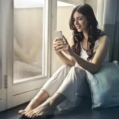 Uśmiechnięta kobieta siedzi przy oknie, przeglądając telefon. Szybki h+ internet pozwala jej cieszyć się chwilą relaksu.