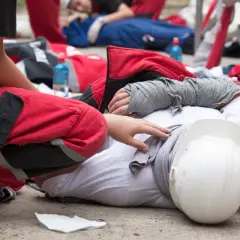 Ratownik w czerwonym stroju ocenia stan rannego, który leży na ziemi z zabandażowaną ręką. To przykład triage po polsku.