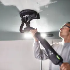 Un homme utilise une ponceuse de plafond Festool, éclairée par une lampe rasante Festool, pour lisser une surface.