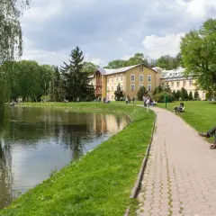 Piękny park przy sanatorium z ZUS. Spacerowicze odpoczywają nad stawem, a w oddali widać żółty budynek.