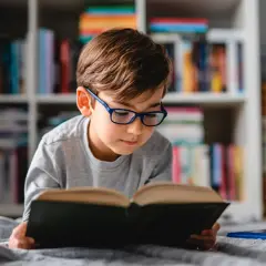 Chłopiec w okularach leży na kocu i czyta książkę na tle regału z książkami.