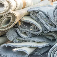 Rotoli di moquette accatastati, pronti per essere rimossi. Si vedono i bordi sfilacciati e i diversi strati dei tappeti.
