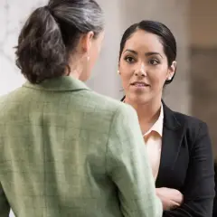 Two women in professional attire converse. One, facing away, wears a green tweed jacket. The other, facing forward, wears a black blazer and light pink blouse, perhaps discussing her northeastern MPA studies.