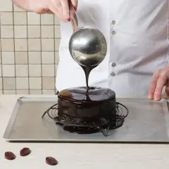 Ein Konditor überzieht einen Kuchen mit glänzender mirror glaze. Schokoladenstückchen liegen daneben.