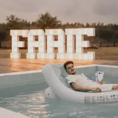 Mężczyzna relaksuje się na dmuchanym materacu w basenie, z wielkimi literami "FARTE" w tle. Idealne miejsce na kawalerski!