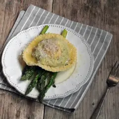 Ravioli z żółtkiem, podane z zielonymi szparagami i posypane parmezanem, na białym talerzu.