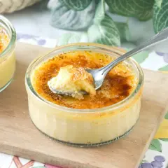 Dos cremosos postres de creme brulee en vasitos de cristal, uno con una cucharada sacada, sobre una tabla de madera.
