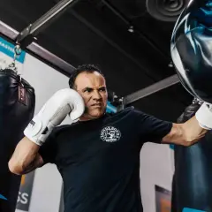 Man with intense focus, performing boxing workouts at home with a heavy bag. He wears white gloves and a black t-shirt.