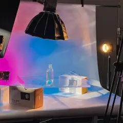 Product photography setup with a bottle and boxes on a table, illuminated by colorful background light and studio lights.