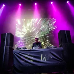 DJ na scenie, fioletowe światła, wizualizacje na ekranie. Sprawdź audioriver timetable, by nie przegapić jego występu!