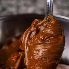 Gładka, brązowa masa czekoladowa na łyżce, w tle metalowa miska.