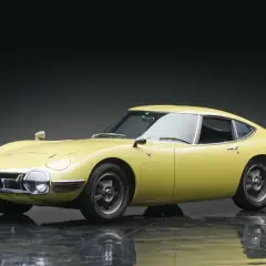 Żółta Toyota 2000 GT z lat 60. prezentuje swoje klasyczne, sportowe linie na błyszczącej podłodze.