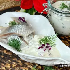Delikatne śledzie w śmietanie z koperkiem i czerwoną cebulką, podane w białej miseczce. Obok słoiczek z sosem.