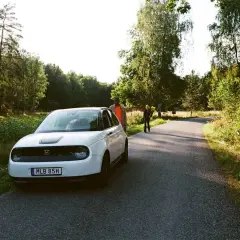 Vit elbil parkerad vid en skogsväg, redo för en bilsemester i Europa. Två personer står vid sidan av vägen.