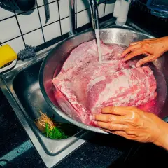 Ręce płuczą duży kawałek wieprzowiny pod bieżącą wodą. To ważny krok, by dowiedzieć się, ile gotować wieprzowinę.