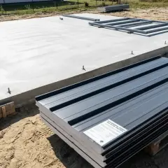 Fundament pod garaż z płyty warstwowej projekt. Betonowa płyta z kotwami i stosem szarych paneli gotowych do montażu.