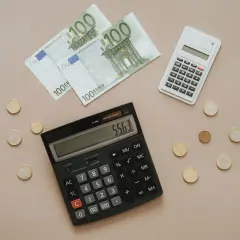 Calcolatrice e banconote da 100 euro, con monete sparse. Si sta calcolando quanto costa la tassa di soggiorno.