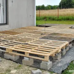 Budowa tarasu z palet. Drewniany podest z palet, podparty na bloczkach, tworzy tymczasową konstrukcję przy domu.