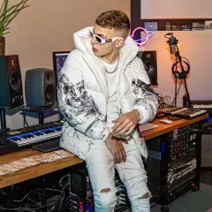 Un giovane skinny cantante con occhiali bianchi e giacca imbottita è seduto in uno studio di registrazione, circondato da tastiere e apparecchiature audio.